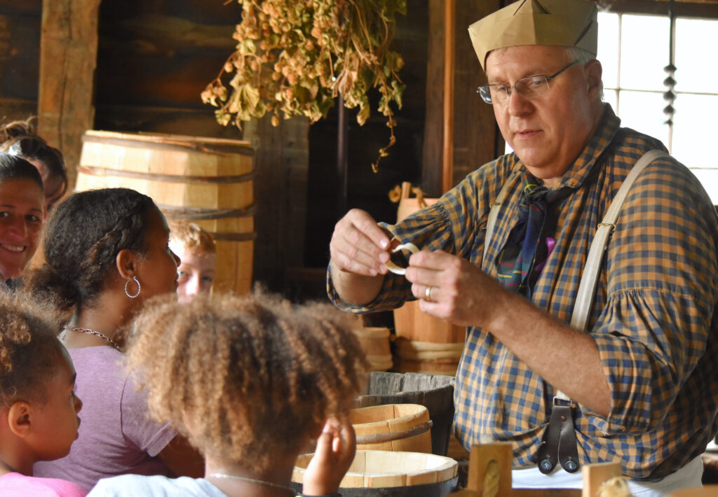 A man in historic costume showing a group of kids what he is working on.