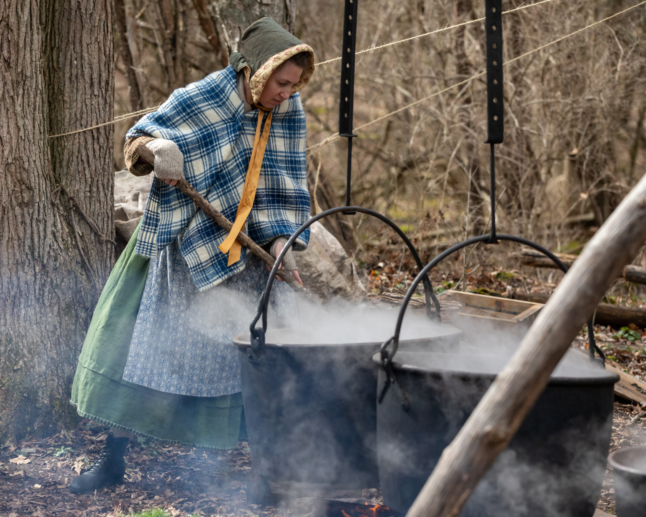 Woman in historic clothing stirring large pots of maple syrup